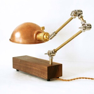 May include: A copper-colored desk lamp with a wooden base and adjustable metal arms. The lamp features a dome-shaped shade, brass accents, and a brown, braided power cord. The lamp's base is rectangular and made of dark wood.