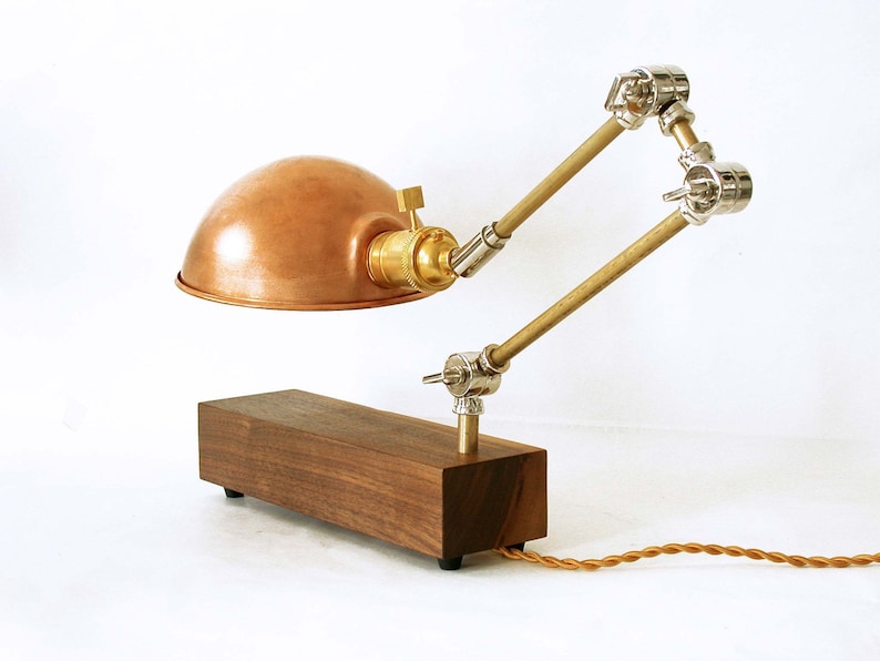 May include: A copper-colored desk lamp with a wooden base and adjustable metal arms. The lamp features a dome-shaped shade, brass accents, and a brown, braided power cord. The lamp's base is rectangular and made of dark wood.
