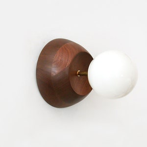Puede incluir: Una lámpara de pared con una base de madera de nogal y un globo de vidrio blanco. La lámpara tiene un acento de latón.