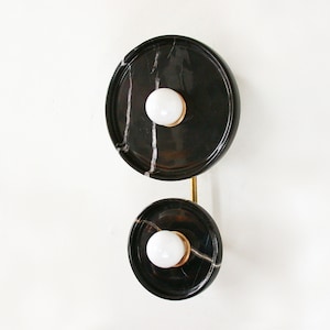 Puede incluir: Una lámpara de pared de mármol negro con dos apliques redondos. Cada aplique tiene una bombilla blanca y una base de metal dorado.