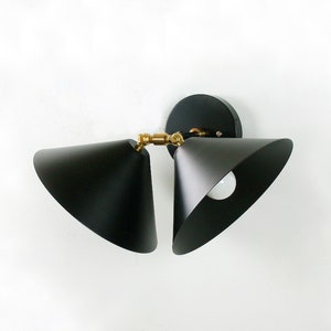 May include: A modern black wall sconce with two cone-shaped shades. The fixture has a circular base and brass-colored joints. The light bulbs are visible inside the shades. The sconce is mounted on a white wall.