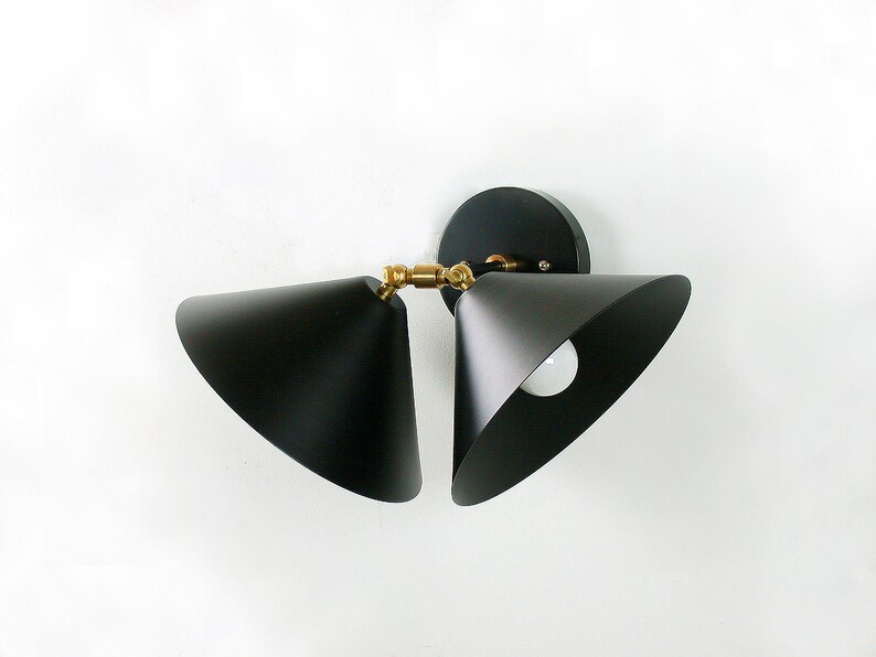May include: A modern black wall sconce with two cone-shaped shades. The fixture has a circular base and brass-colored joints. The light bulbs are visible inside the shades. The sconce is mounted on a white wall.
