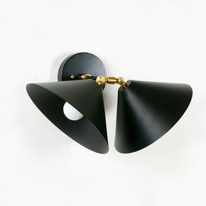May include: Black, cone-shaped wall sconce with a brass-colored joint. The sconce has a circular base and a white light bulb is visible inside the cone. The fixture is mounted on a white wall. Modern lighting fixture.