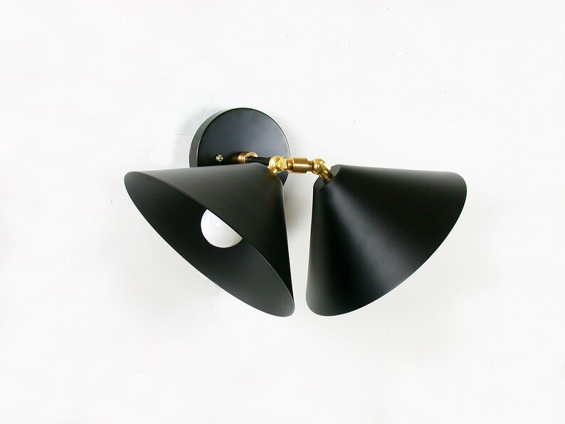 May include: Black, cone-shaped wall sconce with a brass-colored joint. The sconce has a circular base and a white light bulb is visible inside the cone. The fixture is mounted on a white wall. Modern lighting fixture.