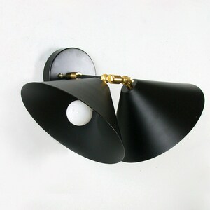 May include: Black wall sconce with two adjustable cone-shaped shades. The fixture has a round base and brass-colored accents. A white light bulb is visible inside one of the shades. The sconce is mounted on a white wall.