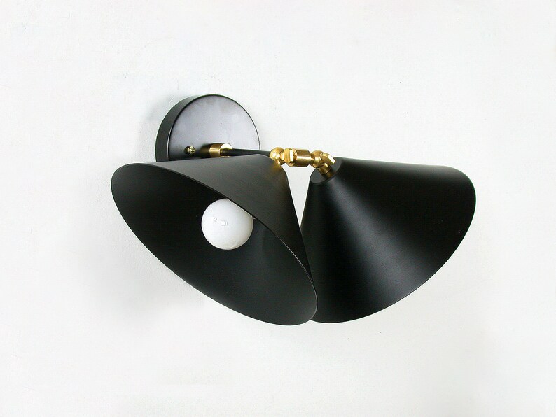 May include: Black wall sconce with two adjustable cone-shaped shades. The fixture has a round base and brass-colored accents. A white light bulb is visible inside one of the shades. The sconce is mounted on a white wall.