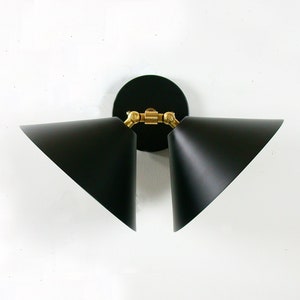 May include: A modern black wall sconce with two triangular shades. The sconce features a circular black base and brass-colored joints. The shades are angled and point outwards, creating a contemporary lighting fixture.