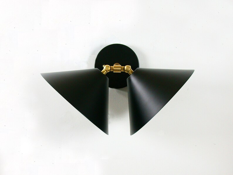 May include: A modern black wall sconce with two triangular shades. The sconce features a circular black base and brass-colored joints. The shades are angled and point outwards, creating a contemporary lighting fixture.