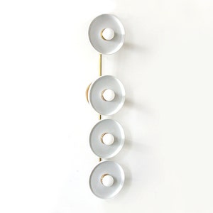 Puede incluir: Una lámpara de pared vertical con cinco luminarias circulares blancas. Cada luminaria tiene un anillo dorado alrededor del borde y una bombilla blanca en el centro.