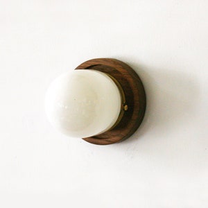 May include: A contemporary wall light with a spherical, white glass shade set in a dark wood base with a brass detail. The light is mounted on a white wall, demonstrating a minimalist aesthetic.