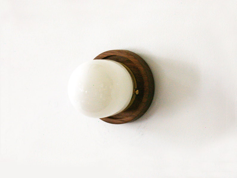 May include: A modern wall sconce featuring a round, white globe light set in a dark wood base with a brass accent. The sconce is mounted on a white wall, showcasing a minimalist design.