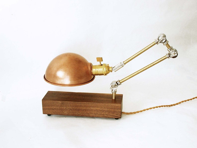 May include: A copper-colored desk lamp with a dome-shaped shade and adjustable brass arms. The lamp sits on a rectangular wooden base and has a vintage-style design. The cord is a twisted brown color.