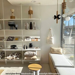 Puede incluir: Una estanter&iacute;a de metal blanco con varios art&iacute;culos de decoraci&oacute;n para el hogar, incluyendo un jarr&oacute;n con estampado en blanco y negro, una escultura de madera y libros. Las estanter&iacute;as est&aacute;n contra una pared blanca con una gran ventana detr&aacute;s de ellas. Un banco blanco con un coj&iacute;n beige se encuentra frente a la ventana.