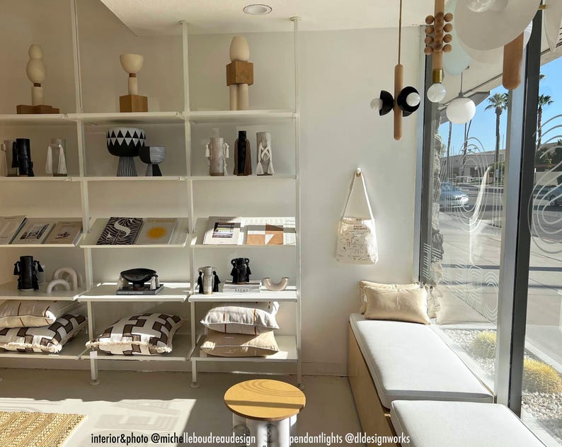 Puede incluir: Una estanter&iacute;a de metal blanco con varios art&iacute;culos de decoraci&oacute;n para el hogar, incluyendo un jarr&oacute;n con estampado en blanco y negro, una escultura de madera y libros. Las estanter&iacute;as est&aacute;n contra una pared blanca con una gran ventana detr&aacute;s de ellas. Un banco blanco con un coj&iacute;n beige se encuentra frente a la ventana.