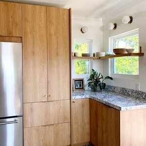K&ouml;nnte beinhalten: Eine K&uuml;che mit Holzschr&auml;nken, einem K&uuml;hlschrank aus Edelstahl und wei&szlig;en Arbeitsplatten. Die K&uuml;che hat einen wei&szlig;en Fliesenboden mit einem schwarz-wei&szlig;en geometrischen Muster. Es gibt zwei Fenster mit wei&szlig;en Rahmen und ein Regal &uuml;ber jedem Fenster. Das Regal &uuml;ber dem Fenster rechts hat ein paar Teller und Schalen darauf. Die K&uuml;che hat ein modernes Design.