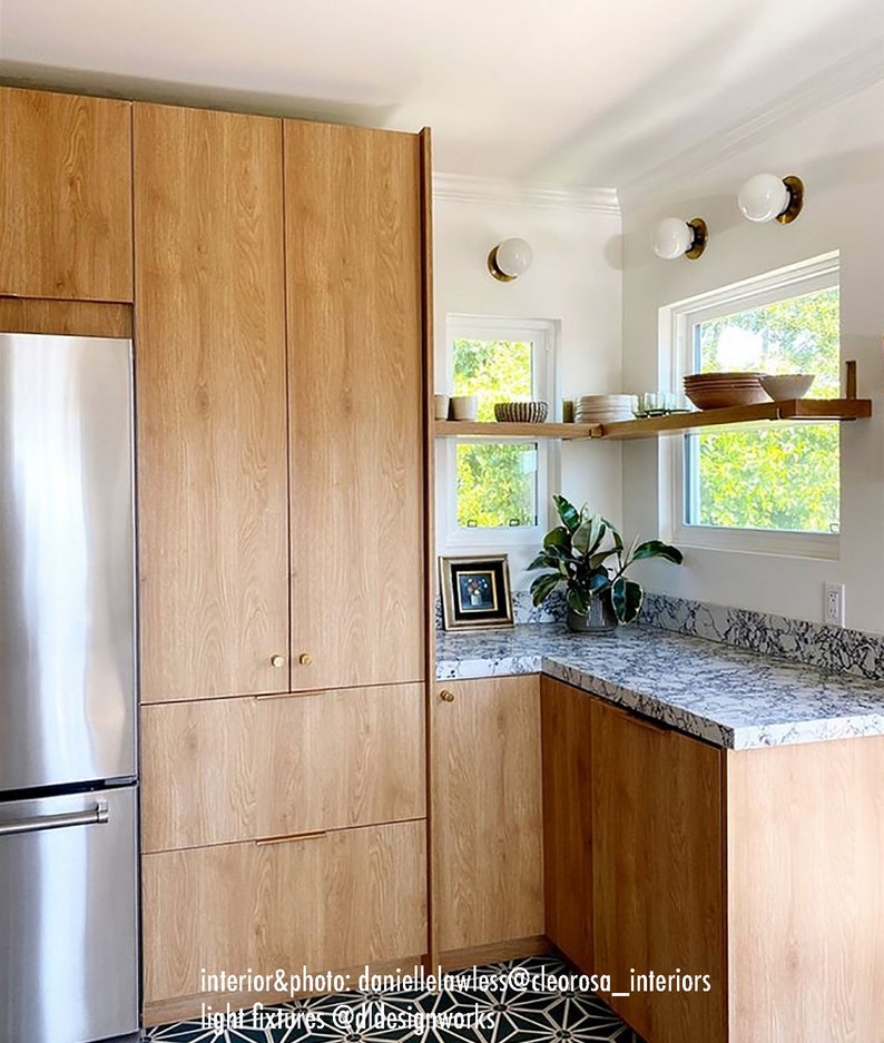 K&ouml;nnte beinhalten: Eine K&uuml;che mit Holzschr&auml;nken, einem K&uuml;hlschrank aus Edelstahl und wei&szlig;en Arbeitsplatten. Die K&uuml;che hat einen wei&szlig;en Fliesenboden mit einem schwarz-wei&szlig;en geometrischen Muster. Es gibt zwei Fenster mit wei&szlig;en Rahmen und ein Regal &uuml;ber jedem Fenster. Das Regal &uuml;ber dem Fenster rechts hat ein paar Teller und Schalen darauf. Die K&uuml;che hat ein modernes Design.