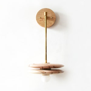 May include: A wooden wall sconce with a brass arm and a double-layered, circular shade. The shade has a natural wood grain pattern.