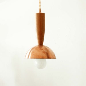 May include: A pendant light fixture featuring a smooth, cylindrical wooden top section connected to a polished copper dome shade. A single round, frosted white light bulb is visible beneath the dome, suspended by a twisted tan fabric cord.