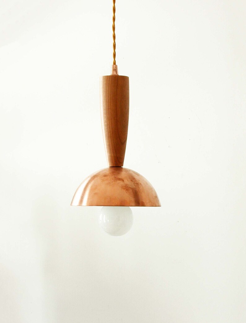 May include: A pendant light fixture featuring a smooth, cylindrical wooden top section connected to a polished copper dome shade. A single round, frosted white light bulb is visible beneath the dome, suspended by a twisted tan fabric cord.