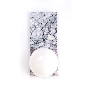 May include: A white marble soap dish with a round, white soap. The soap dish is rectangular with gray veining. The soap is centered on the dish. The background is white.
