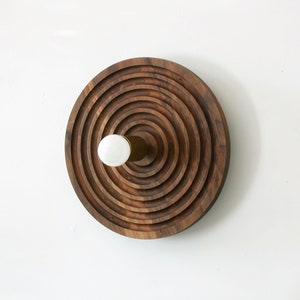 Puede incluir: Una decoración de pared circular de madera con anillos concéntricos de madera, una perilla blanca y una varilla de latón en el centro.