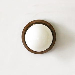 May include: A round, wooden-framed mirror with a white, reflective surface. The dark brown wood frame has a natural grain pattern. The mirror is mounted on a white wall, creating a minimalist aesthetic.