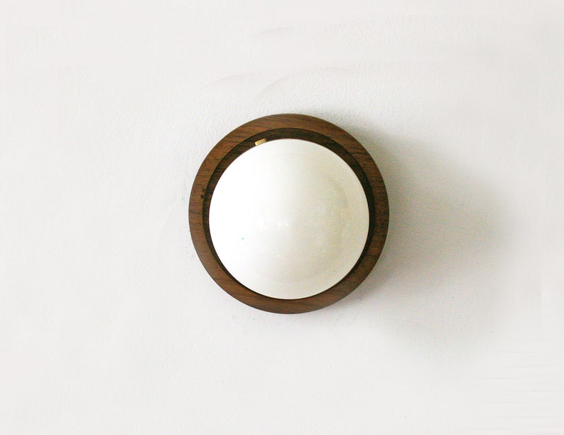 May include: A round, wooden-framed mirror with a white, reflective surface. The dark brown wood frame has a natural grain pattern. The mirror is mounted on a white wall, creating a minimalist aesthetic.