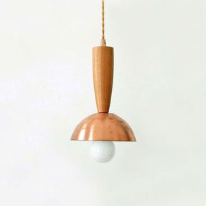 May include: A pendant light fixture featuring a cylindrical light-toned wooden top section connected to a polished copper dome-shaped shade. A single round white light bulb is visible beneath the copper dome, suspended by a twisted fabric cord.