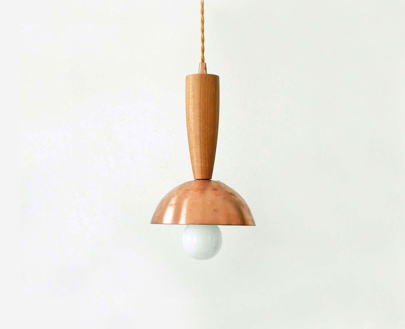 May include: A pendant light fixture featuring a cylindrical light-toned wooden top section connected to a polished copper dome-shaped shade. A single round white light bulb is visible beneath the copper dome, suspended by a twisted fabric cord.
