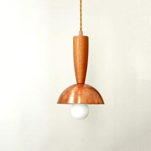 May include: A pendant light fixture featuring a cylindrical light-toned wooden top section connected to a polished copper dome-shaped shade. A single round white light bulb is visible beneath the copper dome, suspended by a twisted tan cord.