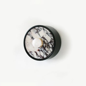 May include: A round, black marble tray with a white marble inlay and a gold knob. The tray is perfect for holding jewelry, keys, or other small items.