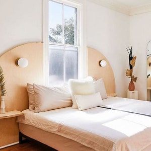 K&ouml;nnte beinhalten: Ein wei&szlig;es Bett mit einem geschwungenen Kopfteil aus hellem Holz. Das Kopfteil hat zwei runde, wei&szlig;e Wandleuchten. Das Bett ist mit wei&szlig;er Bettw&auml;sche und Kissen ausgestattet. Das Bett steht in einem Schlafzimmer mit einer wei&szlig;en Wand und einem Fenster mit wei&szlig;en Rahmen.