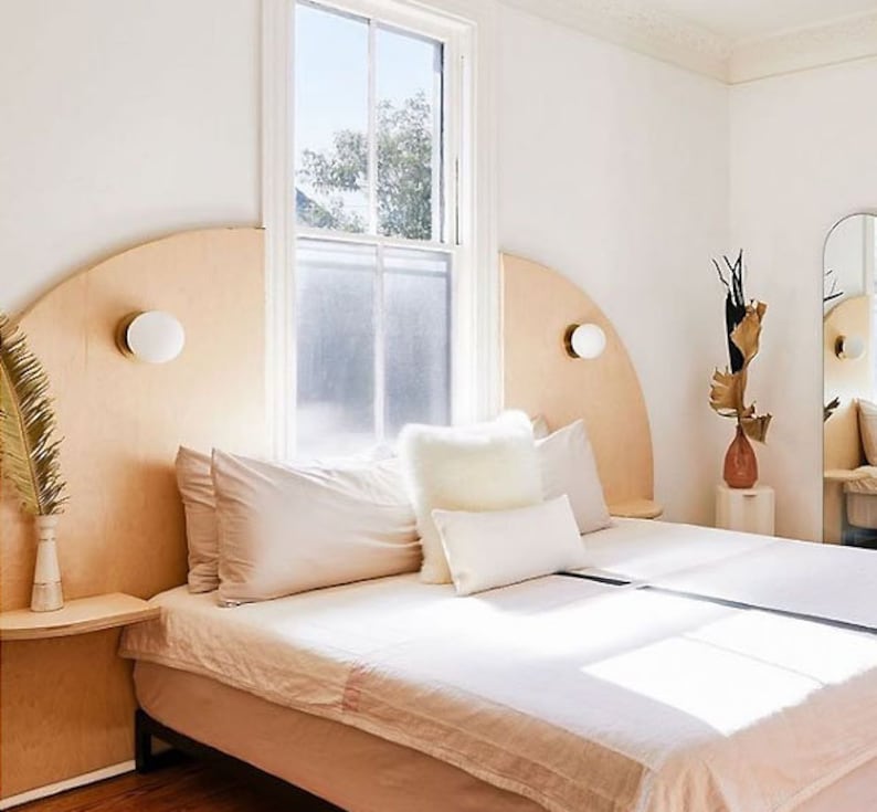 K&ouml;nnte beinhalten: Ein wei&szlig;es Bett mit einem geschwungenen Kopfteil aus hellem Holz. Das Kopfteil hat zwei runde, wei&szlig;e Wandleuchten. Das Bett ist mit wei&szlig;er Bettw&auml;sche und Kissen ausgestattet. Das Bett steht in einem Schlafzimmer mit einer wei&szlig;en Wand und einem Fenster mit wei&szlig;en Rahmen.