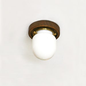 May include: A flush-mount ceiling light fixture with a round, dark wood base and a white, oval-shaped glass shade. The fixture is secured with brass-colored hardware. The light is designed for indoor use.