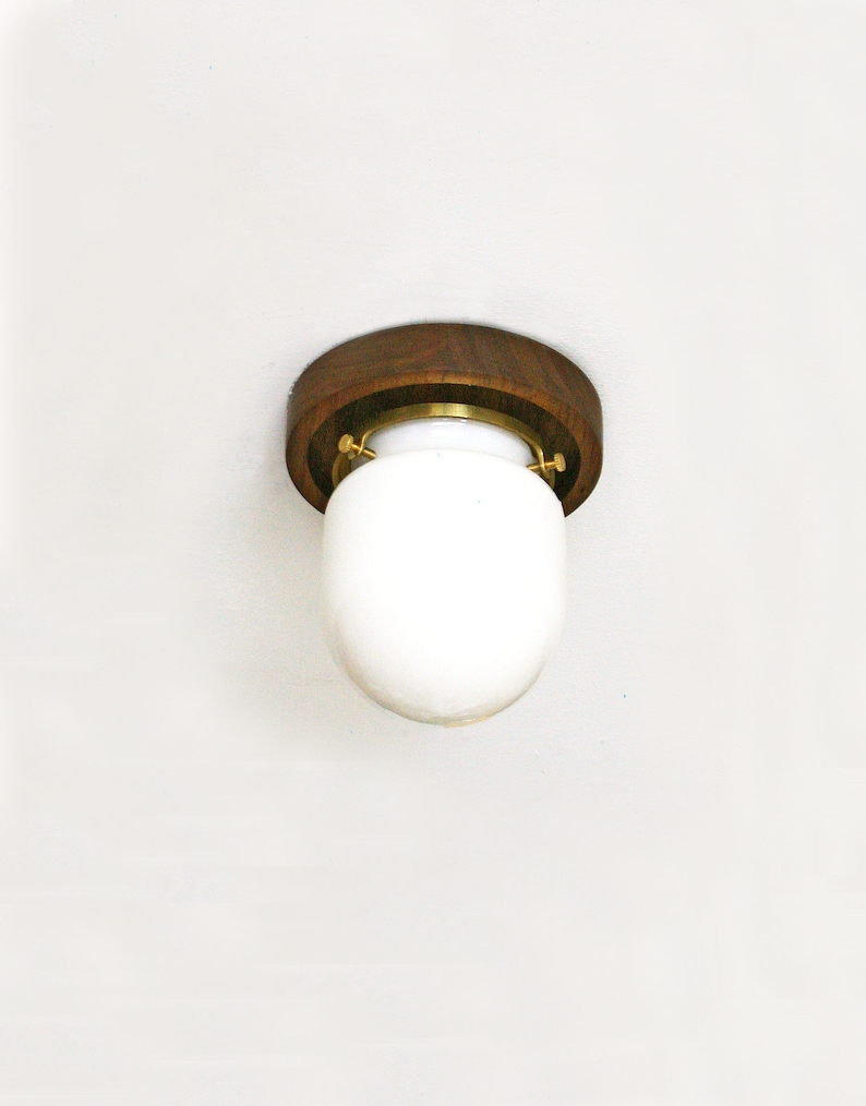 May include: A flush-mount ceiling light fixture with a round, dark wood base and a white, oval-shaped glass shade. The fixture is secured with brass-colored hardware. The light is designed for indoor use.