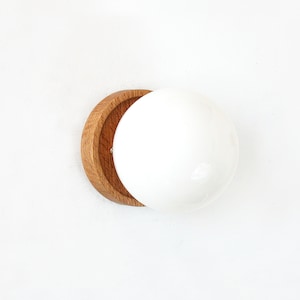 Könnte beinhalten: Eine weiße Keramik-Globus-Leuchte mit einem Holzsockel. Der Sockel ist halbkreisförmig und aus Naturholz gefertigt.