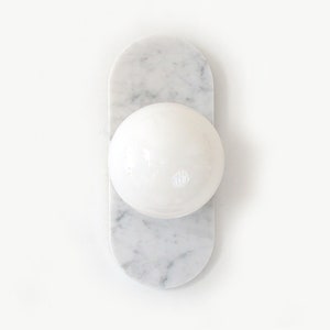 May include: A white marble wall sconce with a rounded, elongated base and a spherical light fixture. The marble has gray veining, and the light is a bright, solid white. The design is minimalist and modern.