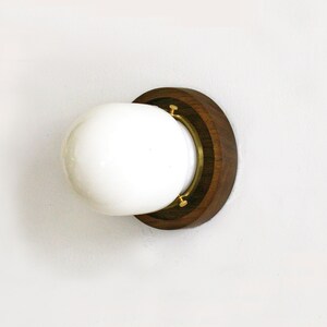 May include: A round, white glass light fixture with a dark brown wooden base and brass accents. The fixture is mounted on a white wall, showcasing a minimalist design. The light fixture is a decorative element.