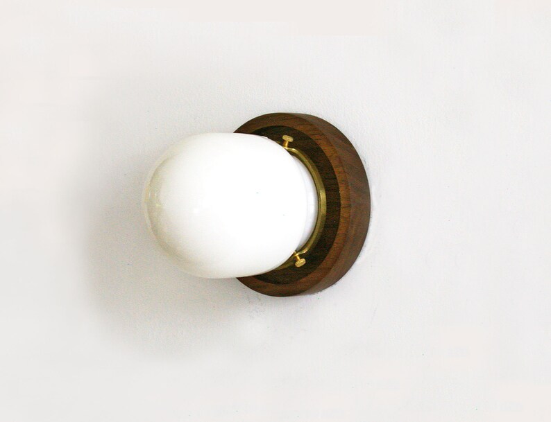 May include: A round, white glass light fixture with a dark brown wooden base and brass accents. The fixture is mounted on a white wall, showcasing a minimalist design. The light fixture is a decorative element.