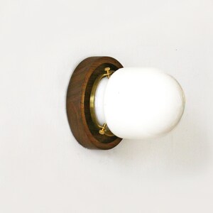 May include: A wall-mounted light fixture featuring a round, white glass globe. The globe is set within a brass ring and a dark brown wooden base. The fixture is designed to be mounted on a white wall.