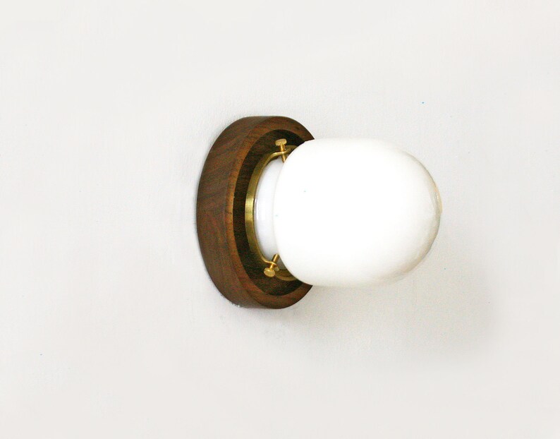 May include: A wall-mounted light fixture featuring a round, white glass globe. The globe is set within a brass ring and a dark brown wooden base. The fixture is designed to be mounted on a white wall.