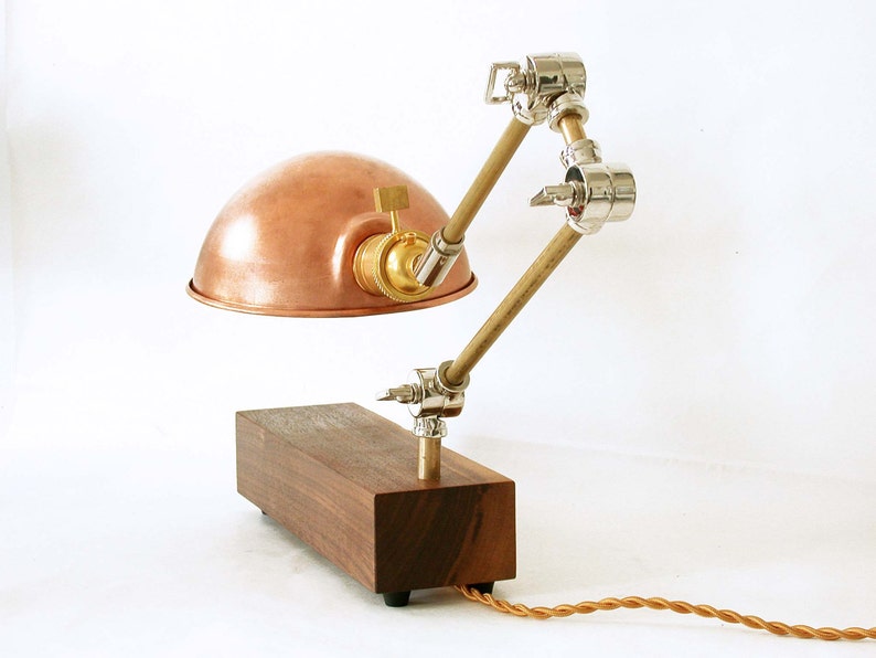 May include: A copper-colored desk lamp with a wooden base and adjustable arm. The lamp features a dome-shaped shade, brass accents, and a brown twisted cord. The adjustable arm is made of wood and metal joints.