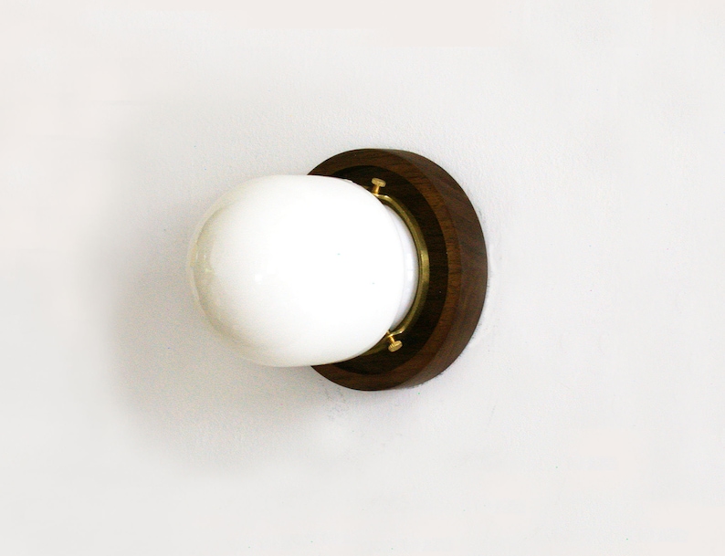 May include: A modern wall sconce featuring a white globe light fixture set within a dark wood and brass circular frame. The sconce is mounted on a white wall, offering a minimalist design suitable for various interior styles.