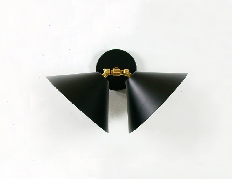 May include: A modern wall sconce with two black, triangular shades. The shades are angled and have white interiors. The fixture has a black circular base and brass-colored joints, creating a stylish lighting element.