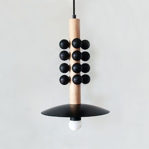 Puede incluir: Una moderna lámpara colgante con una columna central de madera natural, adornada con detalles esféricos negros. Debajo, una pantalla negra en forma de platillo invertido, con una bombilla blanca en la parte inferior. La luz cuelga de un cable negro.