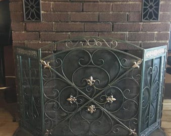 May include: A three-panel fireplace screen made of dark metal with a green-tinted finish. The screen features intricate scrollwork, a central fleur-de-lis motif, and a fine mesh backing. The frame is arched at the top with decorative accents.