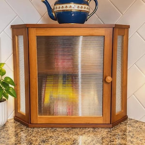 May include: A wooden corner cabinet with a fluted glass door and side panels. The cabinet features a single round wooden knob on the front door. A blue ceramic teapot with a gold rim and a floral pattern sits on top of the cabinet unit.