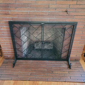 Puede incluir: Una pantalla de chimenea de metal negro con un diseño de dos puertas y un patrón de celosía geométrica. La pantalla está construida con una malla fina y se apoya sobre pies curvos frente a una chimenea de ladrillo rojo.