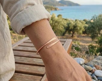 Può includere: Un bracciale a fascia color oro rosa con un design a bypass e due estremità sovrapposte. La superficie metallica presenta una finitura martellata e testurizzata che cattura la luce. Il bracciale è aperto e regolabile sul polso.