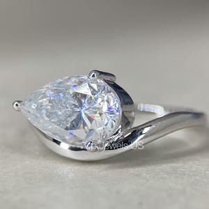 May include: A close-up of a silver ring with a pear-shaped, clear gemstone. The gemstone is the focal point, reflecting light and set in a swirling silver band. The ring is on a neutral background.